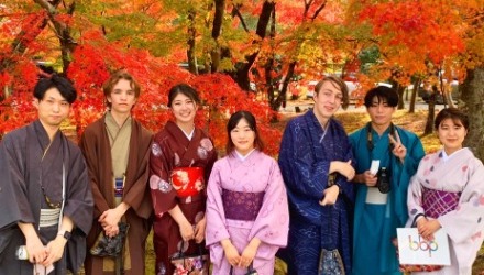 学生7名が秋の着物を羽織り、赤やオレンジや黄色に染まった京都嵐山の美しい紅葉の前でポース
