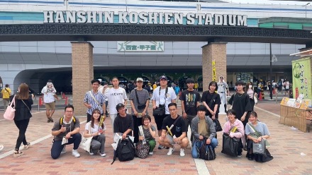 阪神甲子園球場前で15名の学生がタイガーズファングッズのタオルを首に巻いたり、バットを手に持ったりして笑顔で集合写真を撮る